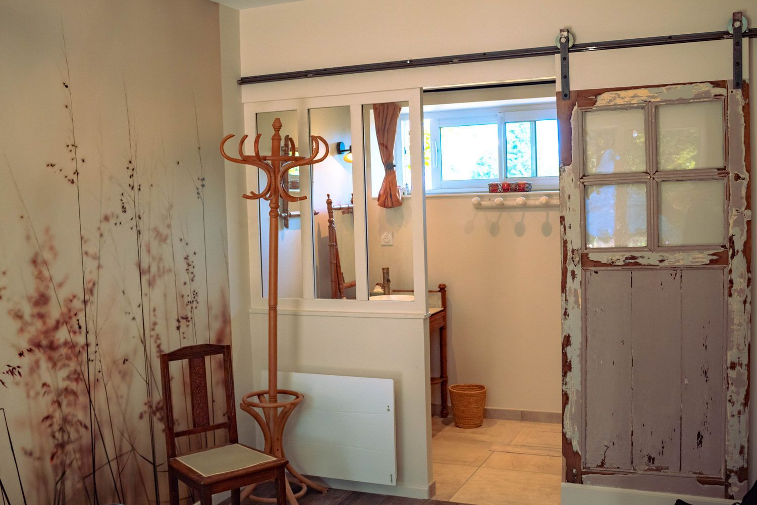 Una habitación rústica con una silla de madera, un perchero, una ventana con cortinas y una puerta con pintura descascarada.