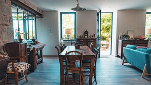 A spacious living area with a wooden dining table, chairs, and a sofa. There is a large window providing natural light and a view of the outdoors. - Les jardins de Nanine (Lapleau)