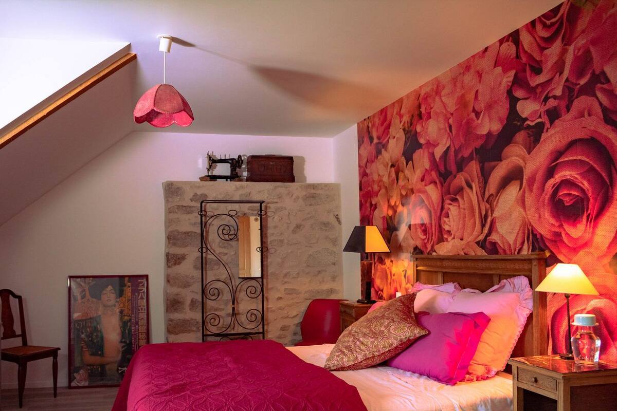 Un dormitorio con techo inclinado, una cama con ropa de cama roja y rosa, un tapiz floral grande en la pared, un cabecero de madera y dos lámparas de noche.