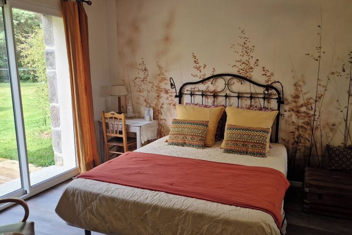 Un dormitorio con cama, una silla de madera, una mesita con lámpara y vistas al exterior.