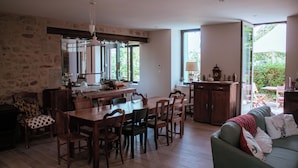 A spacious dining area with a wooden table and chairs, a stone wall, and a view of greenery outside. - Les jardins de Nanine (Lapleau)