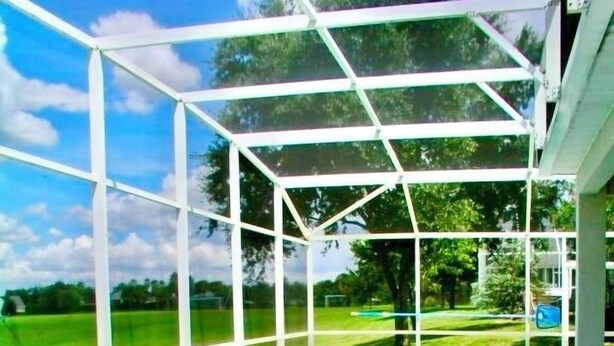Uma piscina com cobertura de vidro, rodeada por um deck branco e vista para um gramado verde.