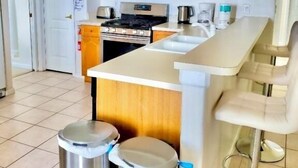 Uma cozinha moderna com armários de madeira, fogão de aço inoxidável e bancada branca. Há duas lixeiras de aço inoxidável e um banquinho de bar.