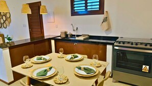 Un coin repas avec une table dressée pour quatre personnes, une cuisine équipée d’une cuisinière et une fenêtre avec des stores.