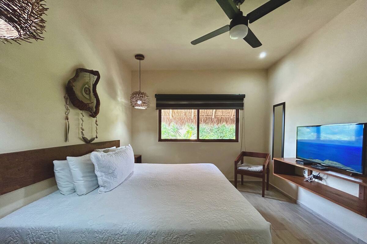 A bedroom with a bed, a ceiling fan, a TV, and a window with a view of greenery.