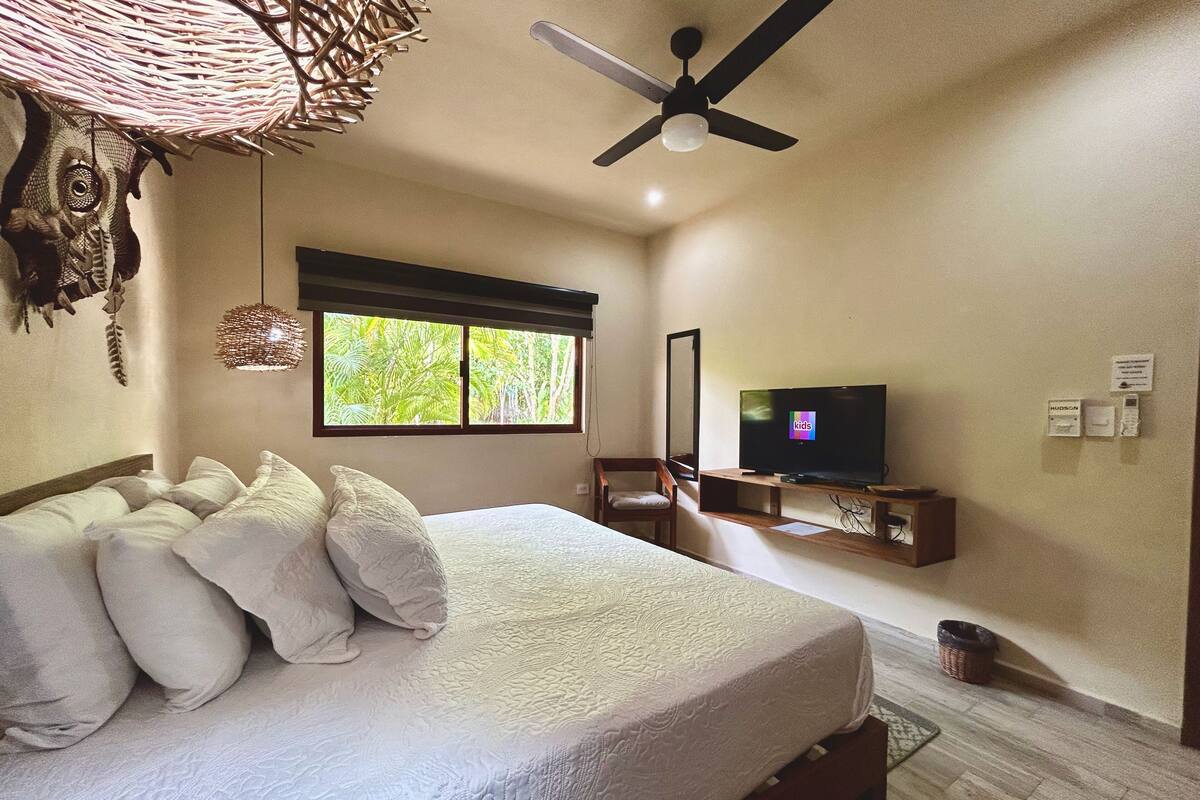 A bedroom with a bed, a ceiling fan, a television, and a window with a view of greenery.