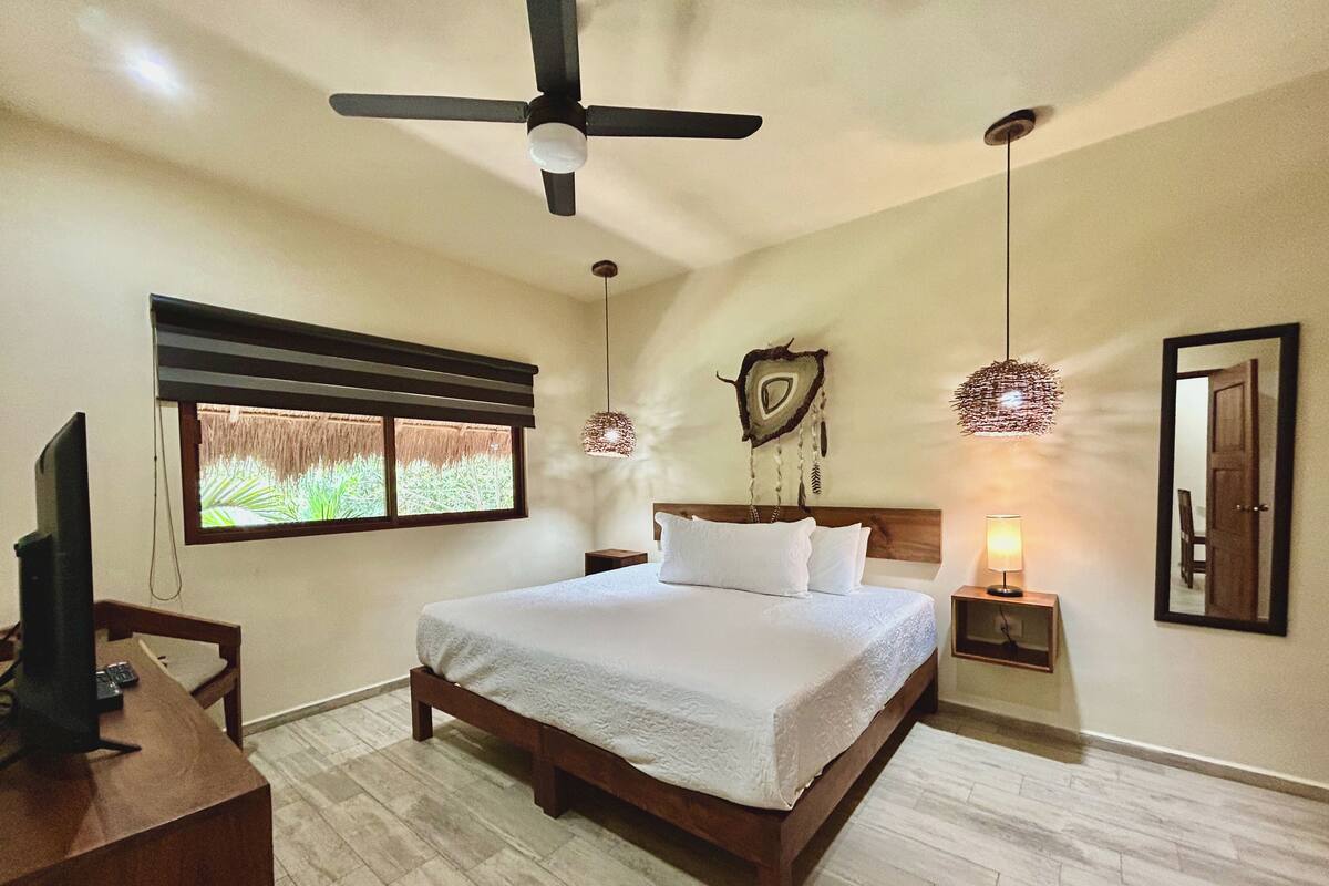 A bedroom with a bed, a ceiling fan, a bedside lamp, a TV, and a window with a view of greenery.