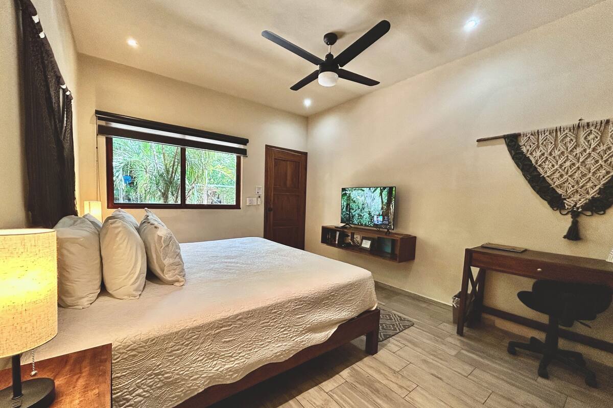 A bedroom with a bed, a ceiling fan, a TV, a desk, and a window with a view of greenery.
