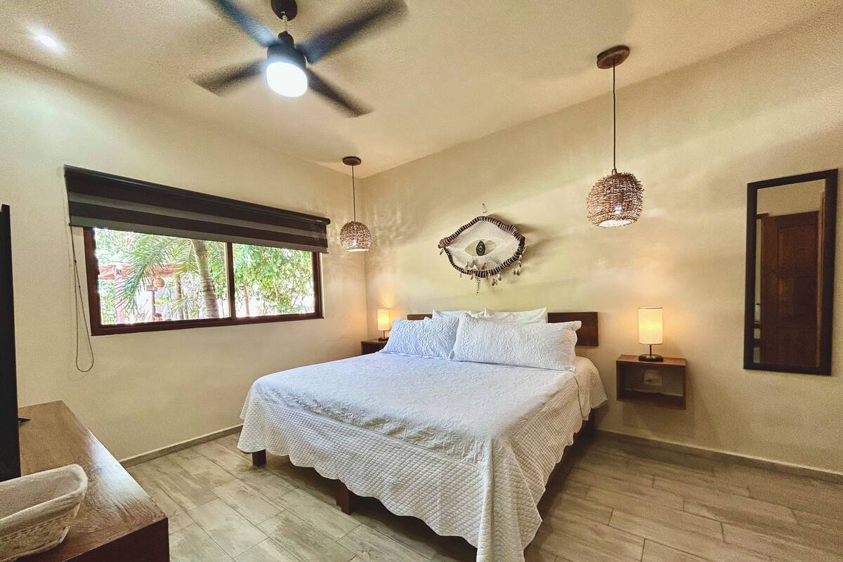 A bedroom with a large bed, two bedside lamps, a ceiling fan, and a window with a view of greenery.