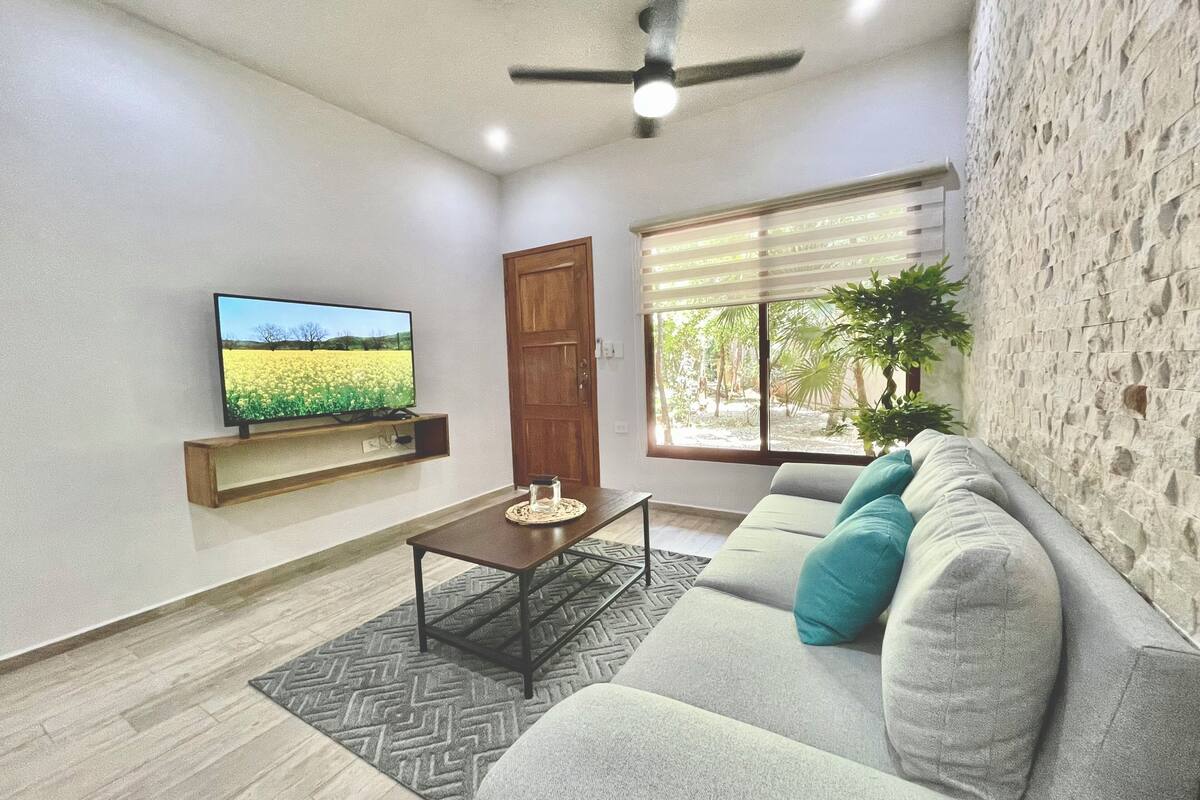 A modern living room with a television, a wooden door, a window with blinds, and a stone wall.