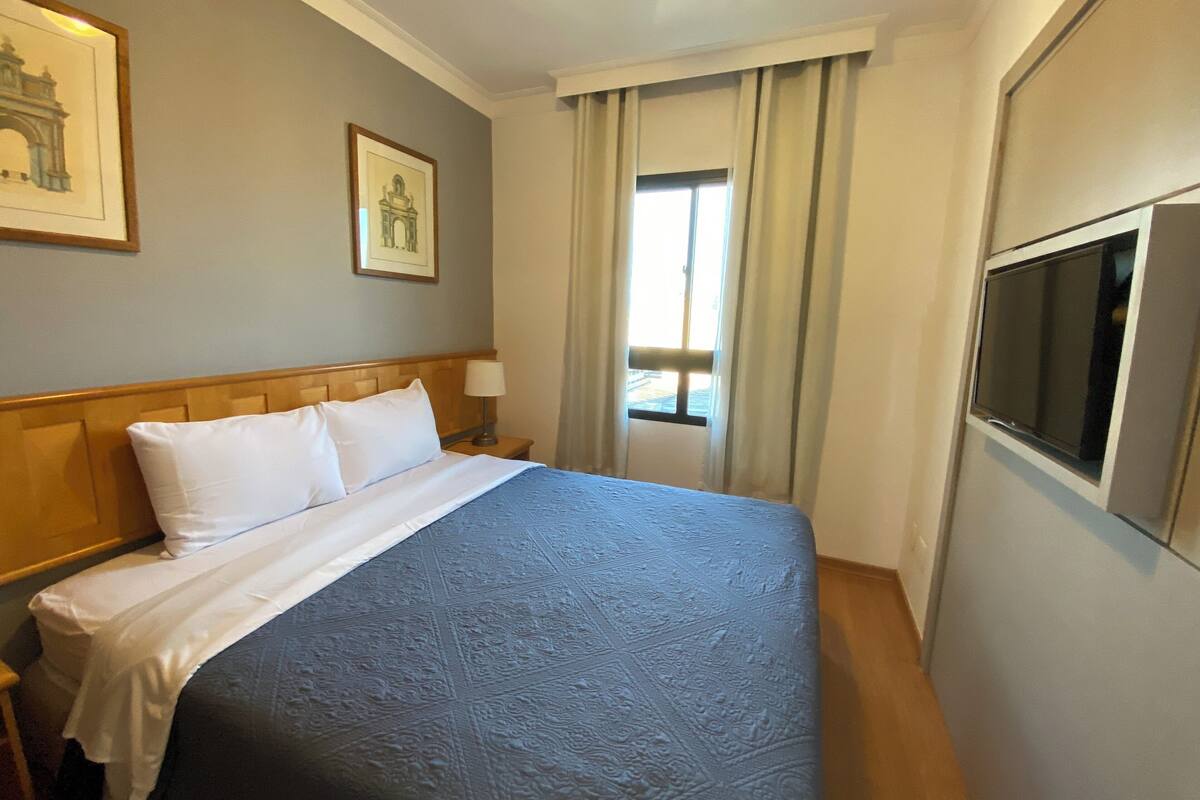 Ein Hotelzimmer mit einem großen Bett, einem an der Wand montierten Fernseher und einem Fenster mit Vorhängen.