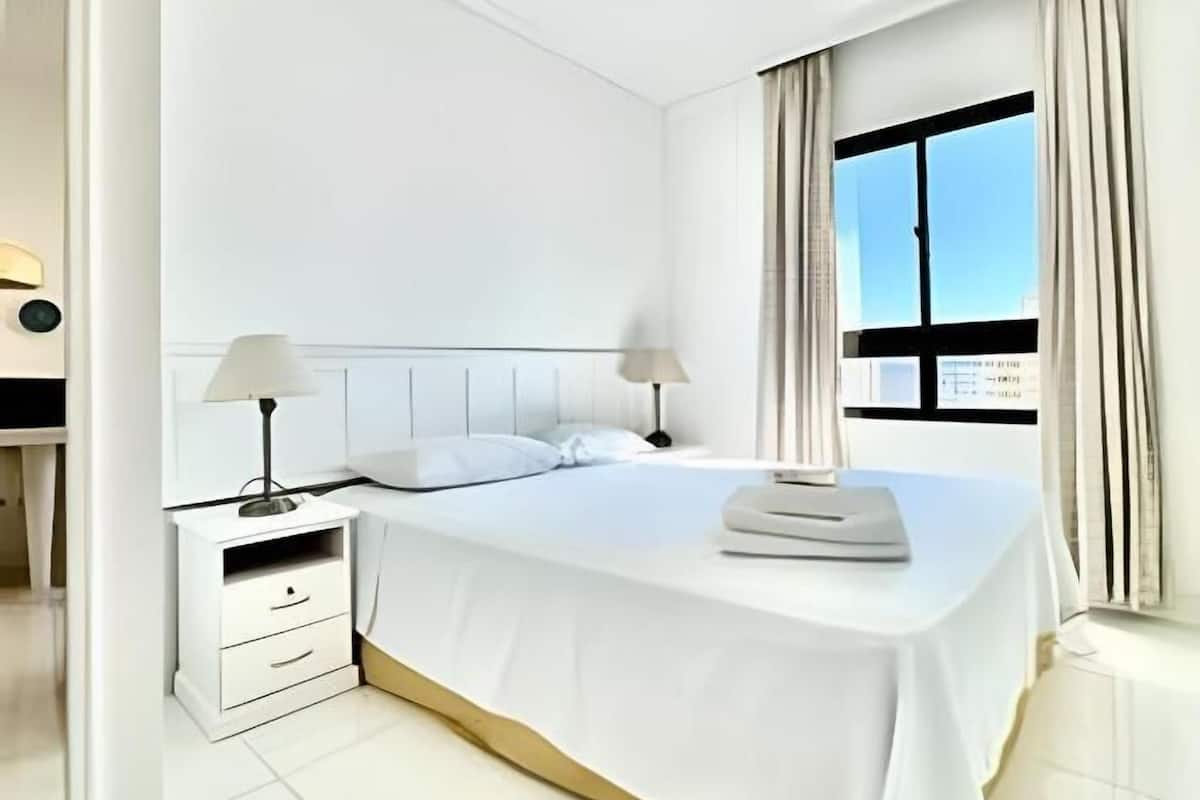 Ein modernes Hotelzimmer mit einem großen Bett, einem Nachttisch mit Lampe, einem kleinen Tisch und einem Fenster mit Vorhängen.