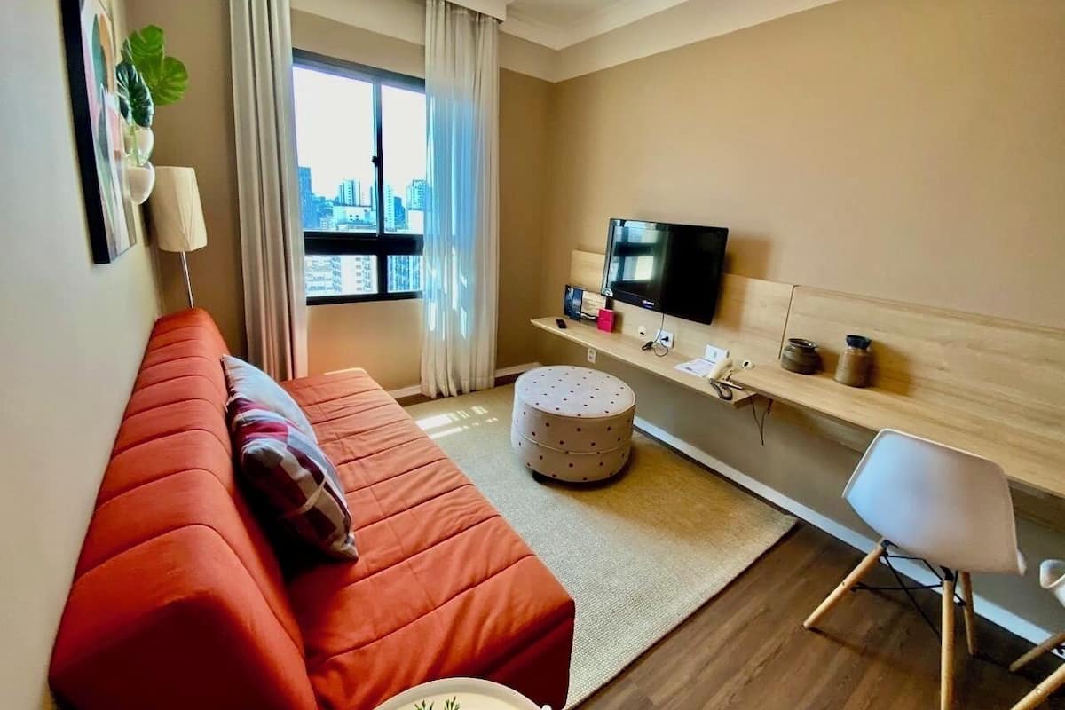 Ein modernes Hotelzimmer mit einem roten Sofa, einem runden Hocker, einem Fernseher auf einem Holzregal und Blick auf die Stadt durch das Fenster.
