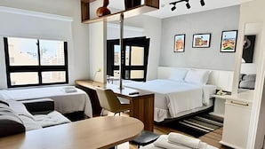 Hotelkamer met twee bedden, een bureau, een stoel en een klein tafeltje. Er hangen afbeeldingen in lijsten aan de muur en een raam met uitzicht op gebouwen.