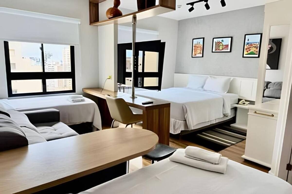 Ein Hotelzimmer mit zwei Betten, einem Schreibtisch, einem Stuhl und einem kleinen Tisch. An den Wänden hängen Bilder und das Fenster bietet einen Blick auf die Stadt.