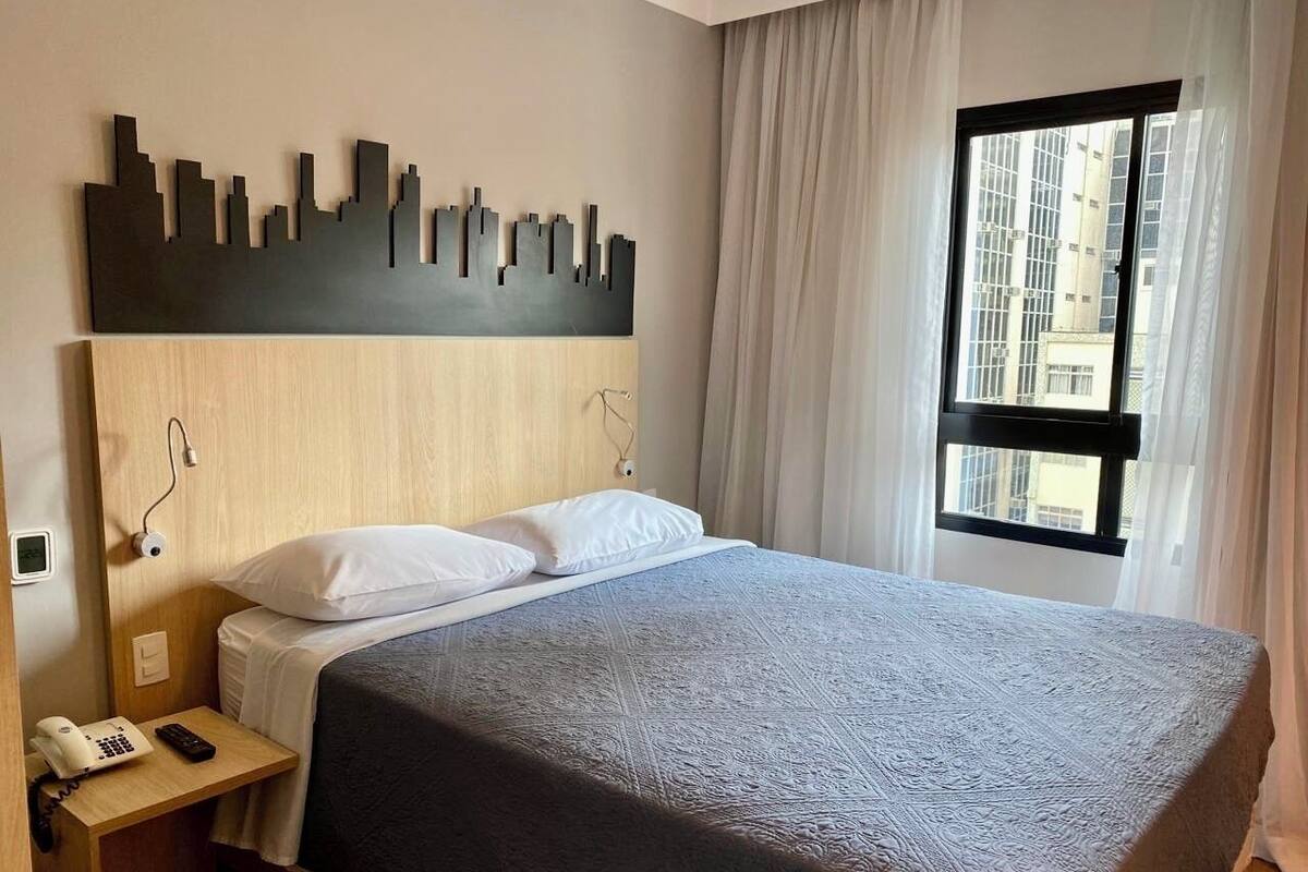 Ein Hotelzimmer mit einem Bett, einem Stadtbild als Kunstwerk, einem Fenster mit Vorhängen und einem Nachttischtelefon.