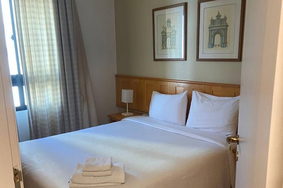 Ein Hotelzimmer mit einem ordentlich bezogenen Bett, einem Nachttisch mit Lampe und zwei gerahmten Bildern an der Wand.