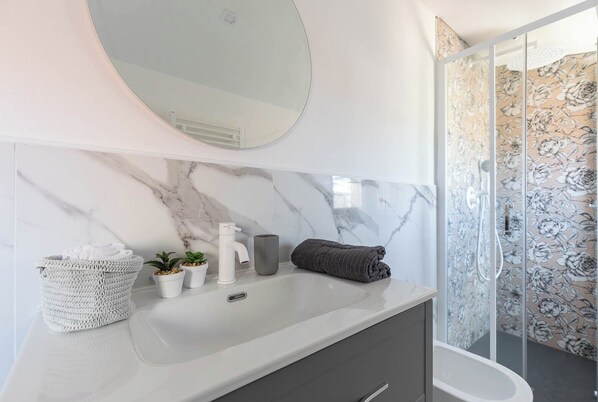 A bathroom with a marble vanity, a round mirror, a shower with floral wallpaper, and a small plant on the counter.