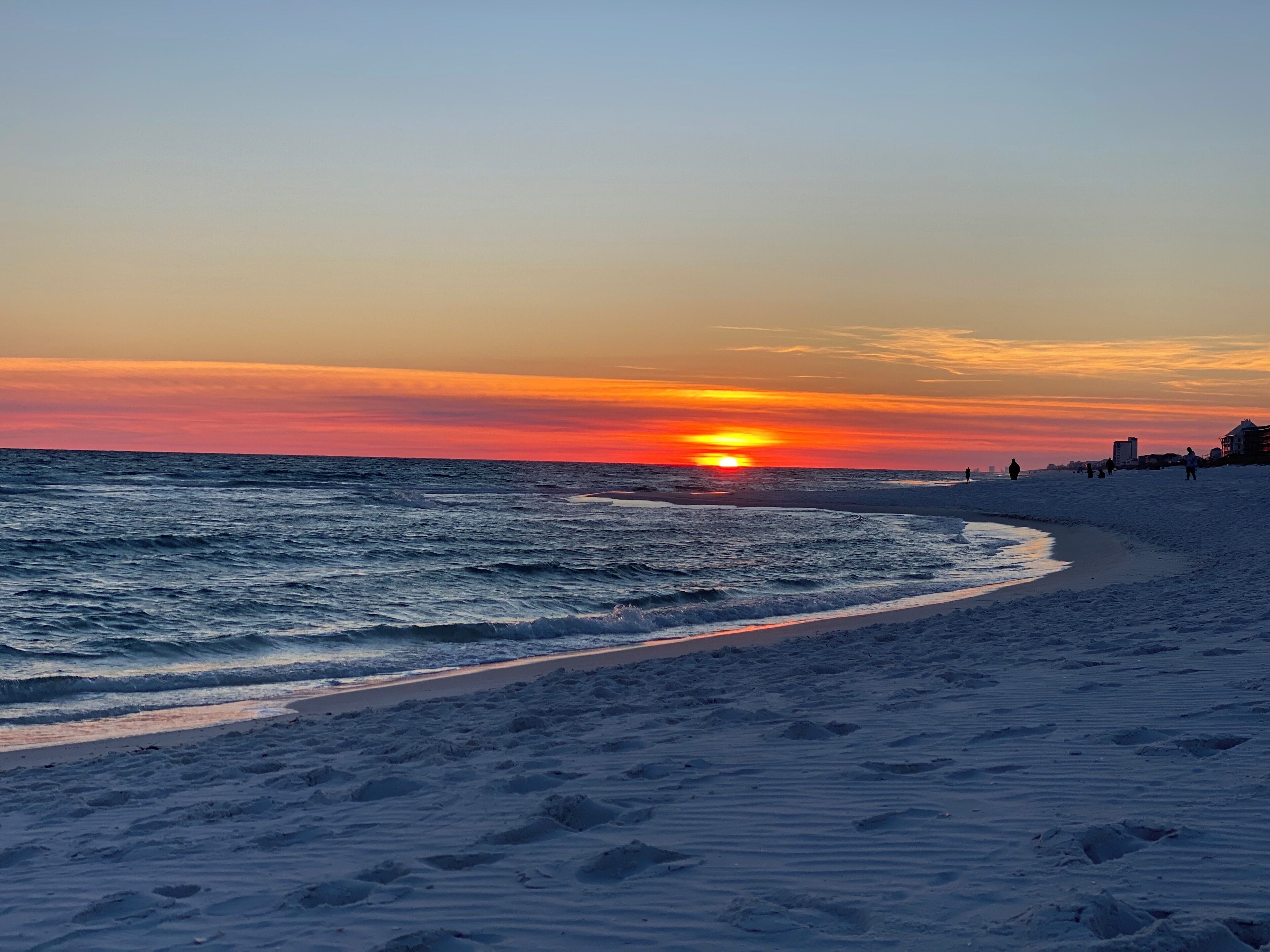 Ein Strand bei Sonnenuntergang mit Wellen, einem sandigen Ufer und Gebäuden im Hintergrund.