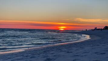 Ein Strand bei Sonnenuntergang mit Wellen, einem sandigen Ufer und Gebäuden im Hintergrund.