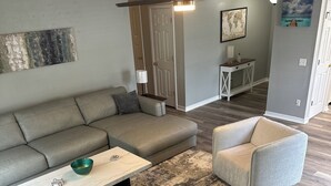 Un salon moderne avec un canapé d’angle, une table basse et deux fauteuils. Le sol est en bois, il y a un tapis à motifs et un ventilateur de plafond.
