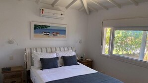 A bedroom with a bed, a window with a view, a painting on the wall, and an air conditioning unit.