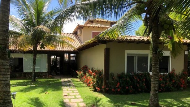 A house with a tiled roof, a stone pathway, and a well-maintained garden with palm trees and red flowers.
