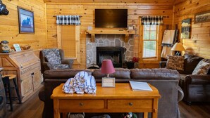 A cozy log cabin interior with a stone fireplace, leather sofas, a wooden table, and a television mounted above the fireplace. - Eagle Eye Cabin in the Smokies (Sevierville)