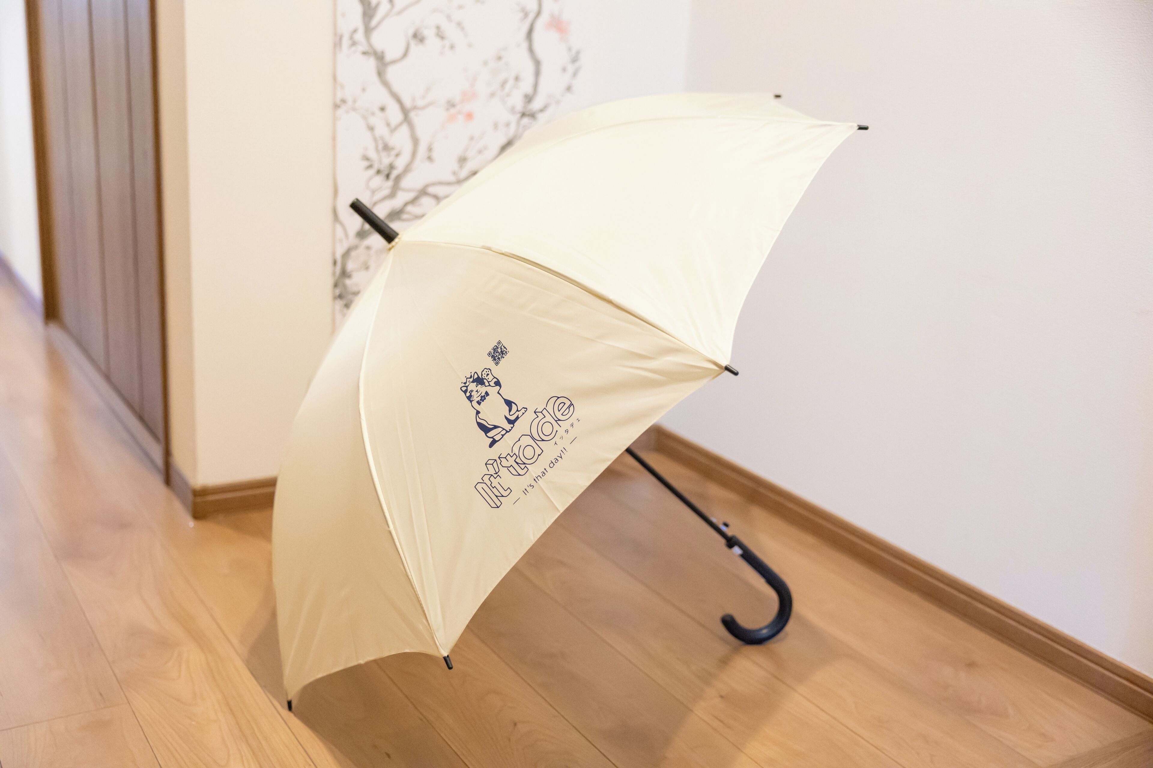 A beige umbrella with a black handle and a logo on the underside, placed on a wooden floor next to a wall with a floral pattern.