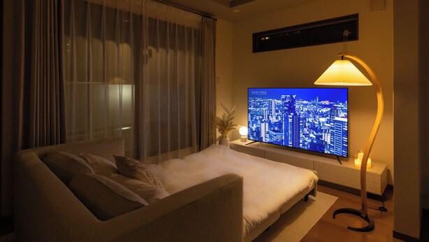 A modern hotel room with a large bed, a flat-screen TV displaying a cityscape, a standing floor lamp, and a window with sheer curtains. - It' ta de ChouChou - Self Check-In Only (Osaka)