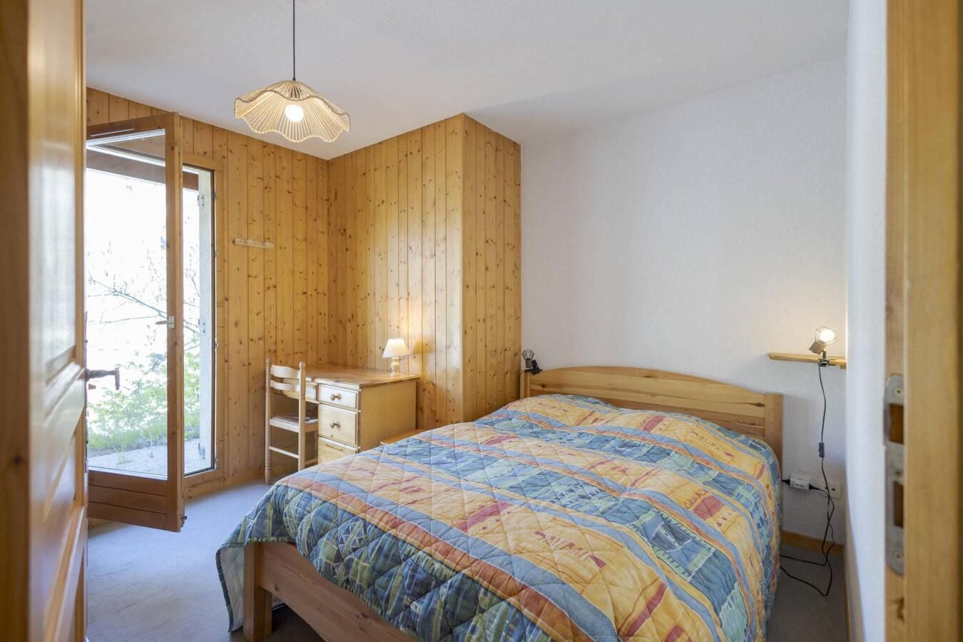 Slaapkamer met een houten bureau, een bed met een kleurrijke sprei, een hanglamp en uitzicht door een open deur.