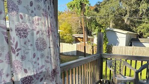 A wooden deck with a railing, a covered area with floral curtains, and a view of a house and trees in the background. - 3-bedroom Mobile Home in enchanting Thunderbolt with WiFi, AC (Thunderbolt)
