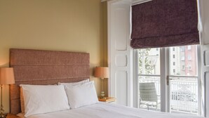 Ein Hotelzimmer mit einem großen Bett, zwei Nachttischlampen, einem Fenster mit Blick auf Gebäude und einem Balkon.