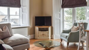 Ein Wohnzimmer mit einer Couch, Sesseln, einem Glastisch, einem Fernseher auf einem Schrank und großen Fenstern mit Vorhängen.