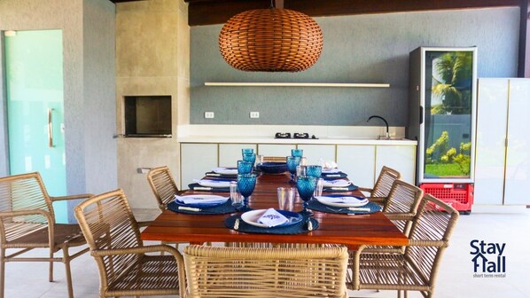 A dining area with a wooden table set for six, surrounded by wicker chairs, a woven pendant light, and a kitchen area with a built-in oven and a large screen displaying a tropical scene.