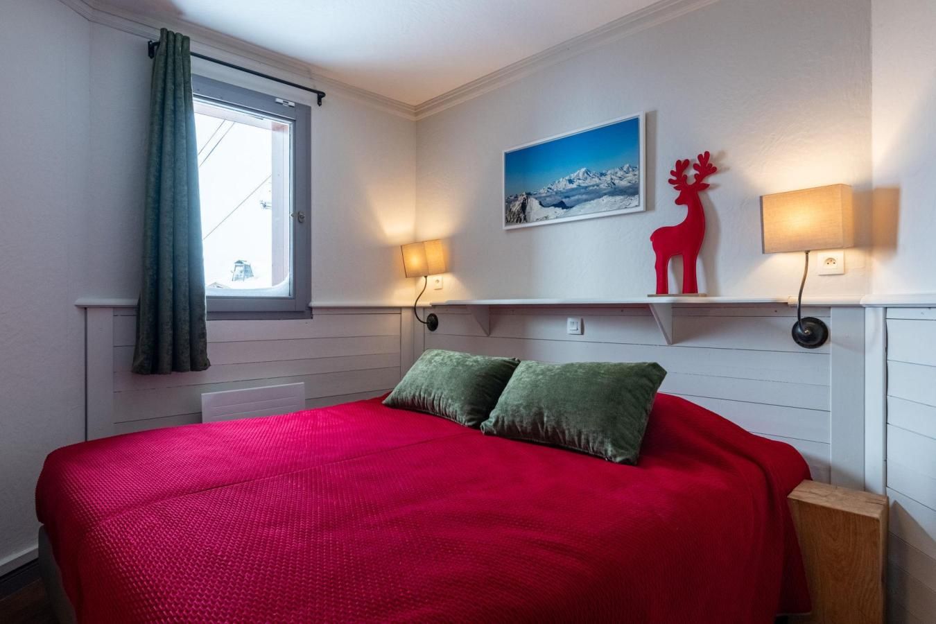 A bedroom with a bed, two pillows, a window with curtains, a wall-mounted shelf with a picture and a reindeer decoration, and two wall-mounted lamps.
