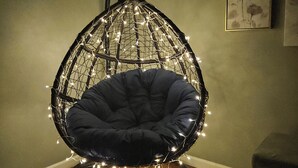 A hanging egg chair with a circular wooden base, surrounded by string lights, in a room with a wooden floor and a gray wall.