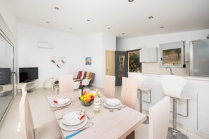 A modern dining area with a table set for two, a television, and a kitchenette with a refrigerator and sink. - Villa Sapfo - Two Bedroom Villa, Sleeps 4 (Lefkada)