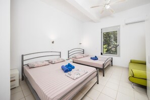 Une chambre avec deux lits, une fenêtre donnant sur des arbres, un ventilateur de plafond et un fauteuil vert.
