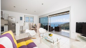 A modern living room with a sofa, dining table, chairs, and a balcony with a view. - Villa Sapfo - Two Bedroom Villa, Sleeps 4 (Lefkada)