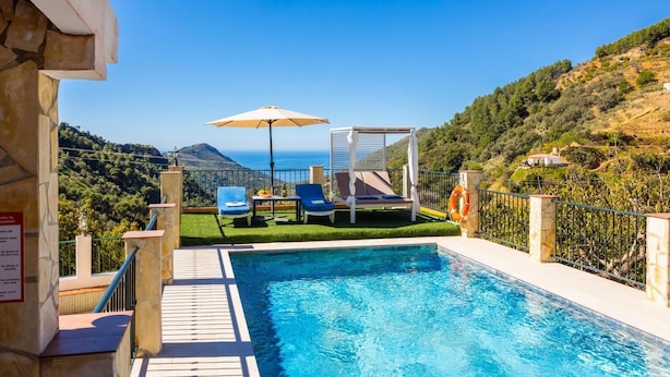 Una piscina con sillones de descanso y una pérgola, rodeada por un muro de piedra y vistas al mar y las colinas.
