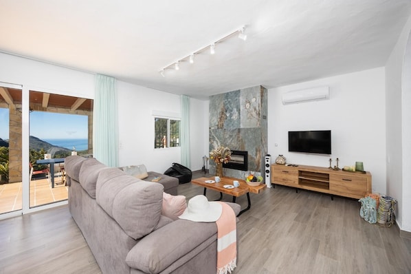 A modern living room with a sofa, a coffee table, a fireplace, a TV, and a balcony with a view.