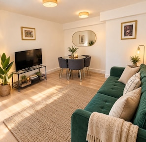 A living room with a green sofa, a TV on a stand, a dining table with chairs, and a mirror on the wall.