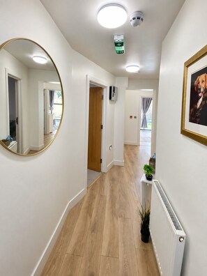 Un couloir doté d’un plancher en bois, de murs blancs, d’un miroir rond, d’un tableau encadré, d’un radiateur et d’un détecteur de fumée fixé au plafond.