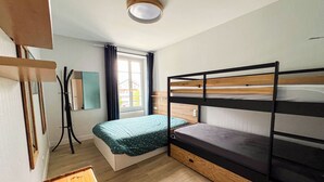 A compact room with a bunk bed, a desk, a chair, a window with curtains, and a radiator. - Spacious T2 in the heart of Chamonix (Chamonix-Mont-Blanc)