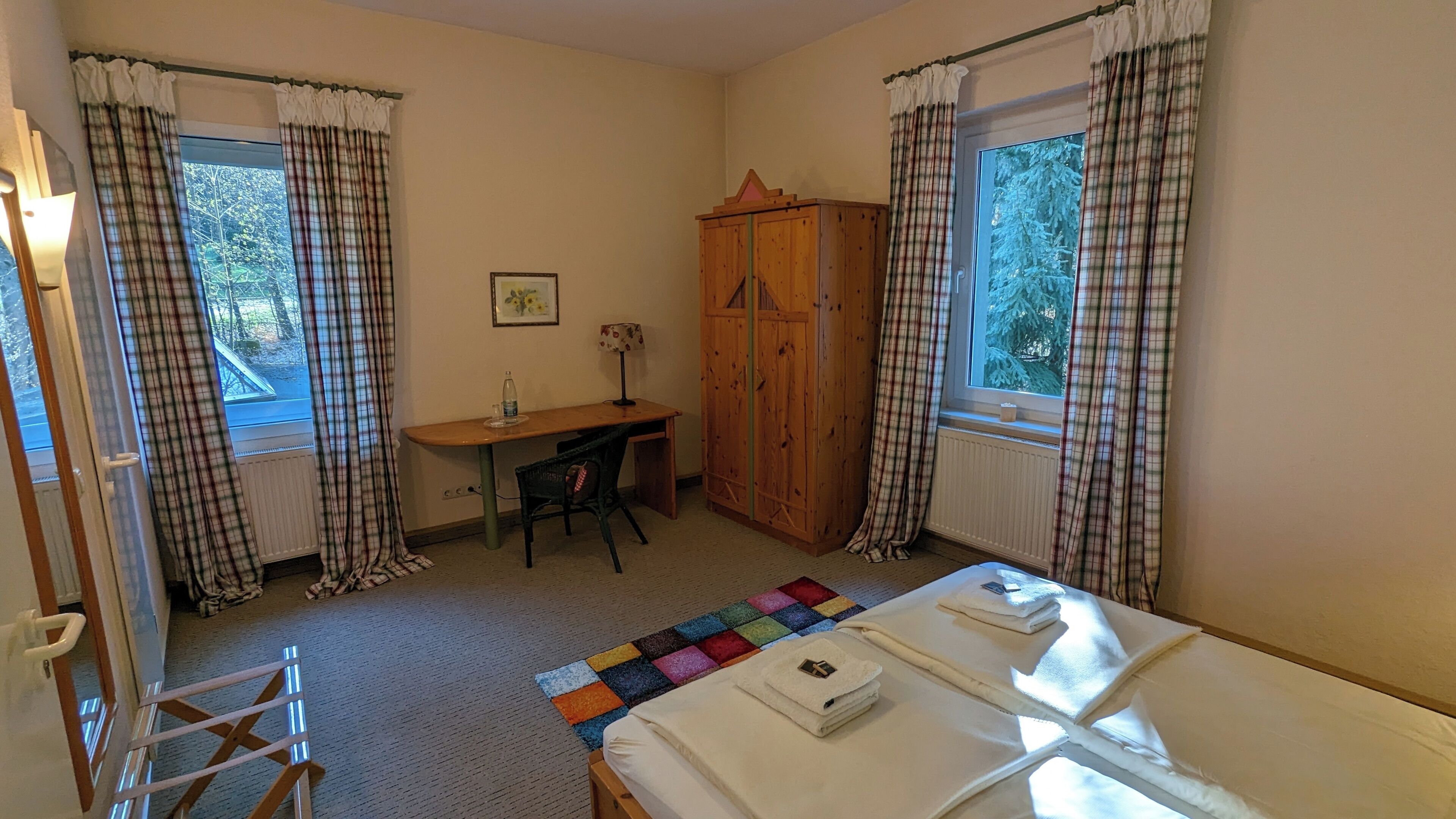 A room with a bed, a desk, a chair, a wardrobe, and two windows with curtains.