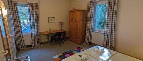 Ein Zimmer mit einem Bett, einem Schreibtisch, einem Stuhl, einem Kleiderschrank und zwei mit Vorhängen versehenen Fenstern.