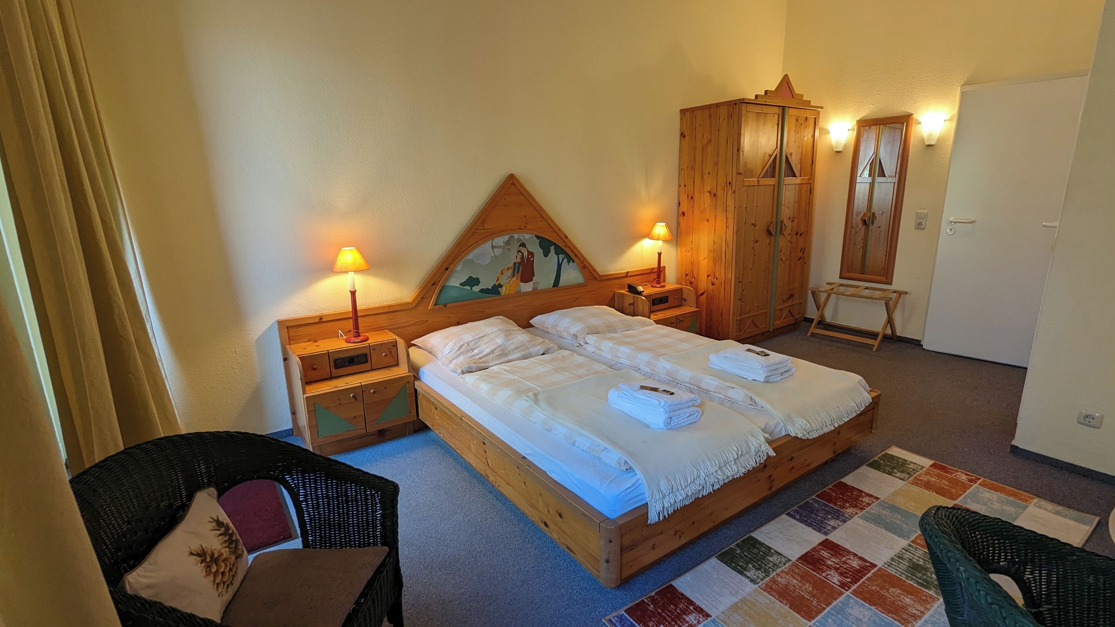 A double bed with two pillows, a wooden headboard, two bedside tables with lamps, a wardrobe, a mirror, and a small chair.