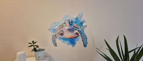 Una pared con una ilustración de tortuga, una pequeña planta en maceta y una caja blanca con un objeto cilíndrico encima.