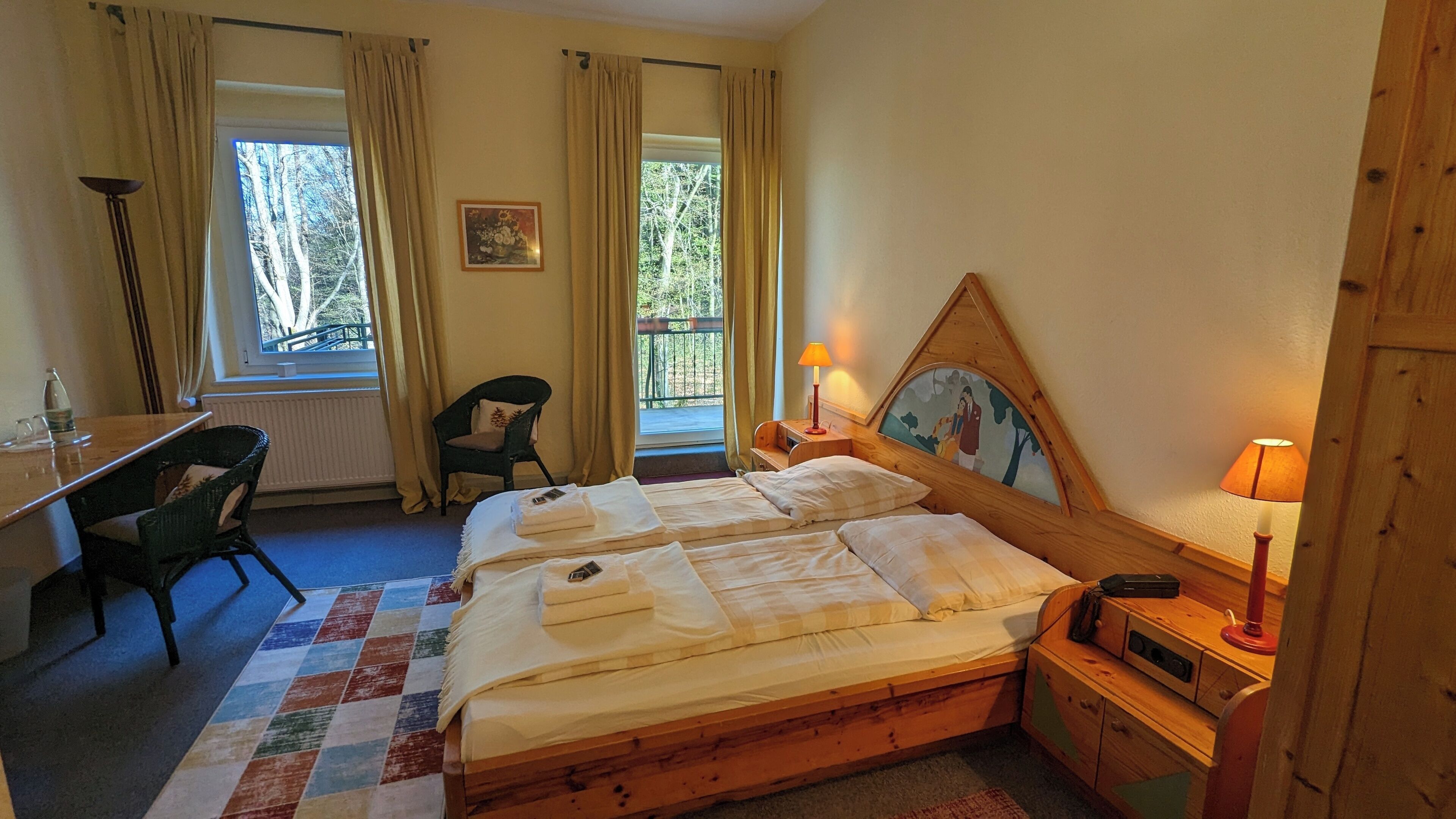 Quarto com duas camas, uma escrivaninha, uma cadeira, janela com cortinas, abajur e um quadro na parede.